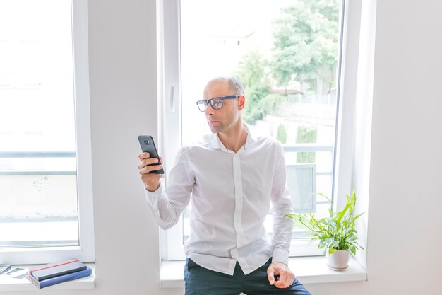 Hombre de negocios sentado frente a la ventana de la oficina con teléfono móvil