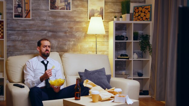 Hombre de negocios sentado en el sofá comiendo patatas fritas comiendo televisión. Zoom en tiro.