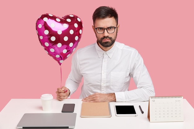 Foto gratuita hombre de negocios serio recibe san valentín de su novia en el lugar de trabajo, sostiene un globo en forma de corazón, posa en el escritorio