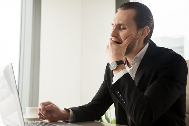 Hombre de negocios soñoliento cansado que bosteza delante de la computadora portátil