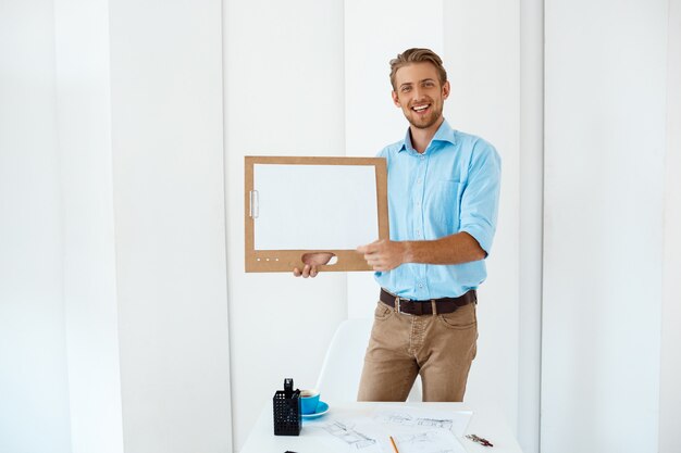 Hombre de negocios sonriente alegre hermoso joven que se coloca en la tabla que sostiene el tablero de madera con la hoja blanca. Luz interior de oficina moderna