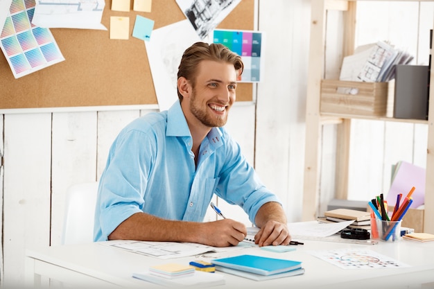 Hombre de negocios sonriente confidente hermoso joven de trabajo que se sienta en la escritura de la tabla en cuaderno. Interior de oficina moderno blanco