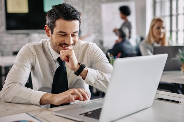 Un hombre de negocios sonriente sentado en un escritorio en la oficina y trabajando en una laptop Hay gente en el fondo