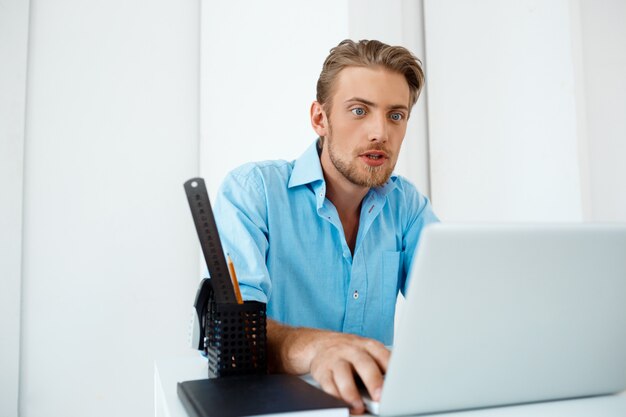 Hombre de negocios sorprendido confiado hermoso joven que se sienta en la tabla que trabaja en la computadora portátil con la taza de café a un lado. Interior de oficina moderno blanco