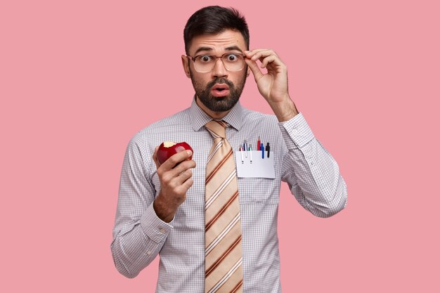 Hombre de negocios sorprendido vestido con camisa formal y corbata, come deliciosa manzana, mira a través de espectáculos con desconcierto