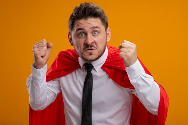Hombre de negocios de superhéroe en capa roja apretando los puños con cara enojada de pie sobre la pared naranja
