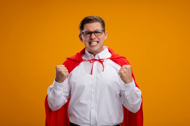 Hombre de negocios de superhéroe en capa roja y gafas apretando los puños con cara enojada volviéndose salvaje parado sobre la pared naranja