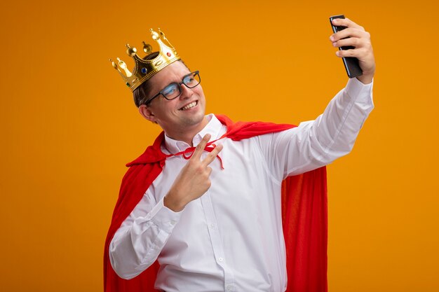 Hombre de negocios de superhéroe en capa roja y gafas con corona haciendo selfie con smartphone sonriendo mostrando signo v parado sobre pared naranja