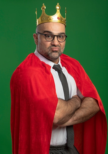 Foto gratuita hombre de negocios de superhéroe en capa roja y gafas con corona mirando al frente sintiéndose orgulloso de sí mismo satisfecho con los brazos cruzados en el pecho de pie sobre la pared verde