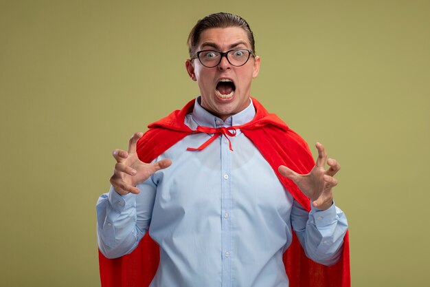 Hombre de negocios de superhéroe en capa roja y gafas gritando con las manos levantadas loco loco volviendo loco de pie sobre fondo claro
