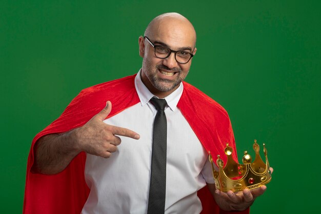 Foto gratuita hombre de negocios de superhéroe en capa roja y gafas sosteniendo corona mirando seguro apuntando sonriente con el dedo índice en la corona sonriendo de pie sobre la pared verde