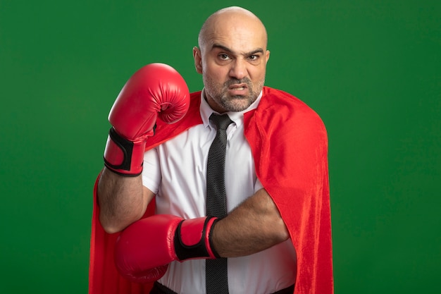 Hombre de negocios de superhéroe en capa roja y guantes de boxeo mirando al frente confundido y disgustado con la mano levantada de pie sobre la pared de luz