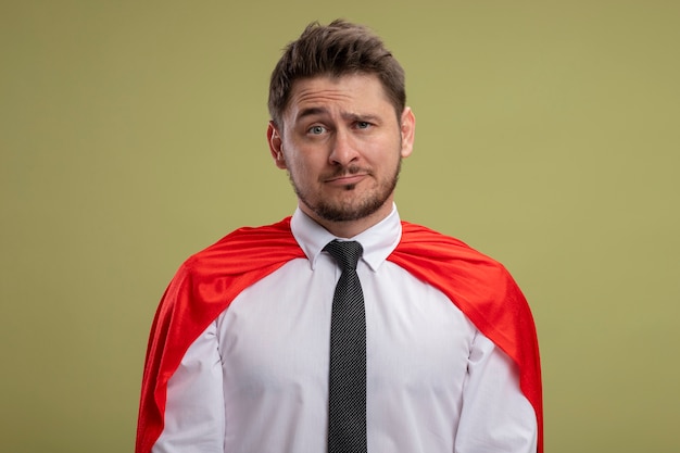 Hombre de negocios de superhéroe en capa roja mirando con expresión triste de pie sobre la pared verde