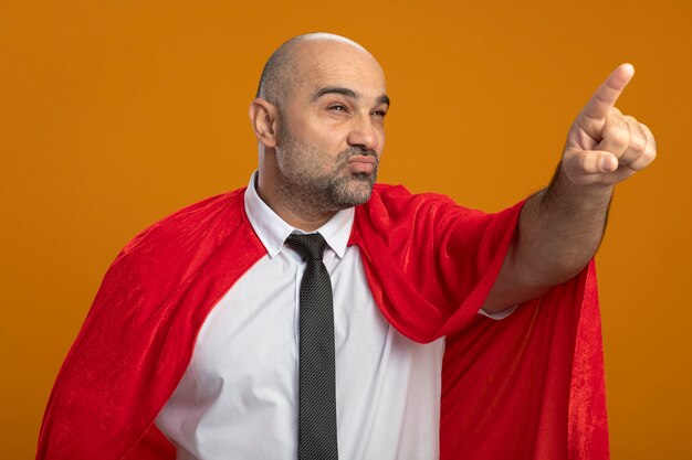 Hombre de negocios de superhéroe en capa roja mirando a un lado con expresión escéptica apuntando con el dedo índice a algo parado sobre la pared naranja