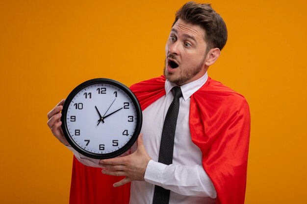 Hombre de negocios de superhéroe en capa roja sosteniendo el reloj de pared mirando asombrado y sorprendido de pie sobre la pared naranja