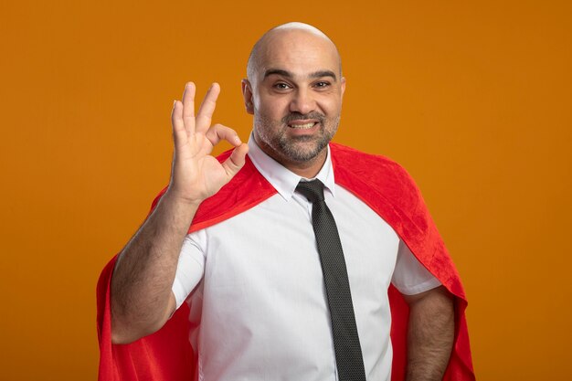 Hombre de negocios de superhéroe complacido en capa roja mirando al frente sonriendo ampliamente mostrando el signo de ok de pie sobre la pared naranja