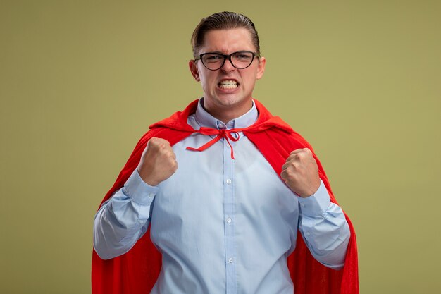 Hombre de negocios de superhéroe enojado en capa roja y gafas apretando los puños con expresión agresiva de pie sobre fondo claro
