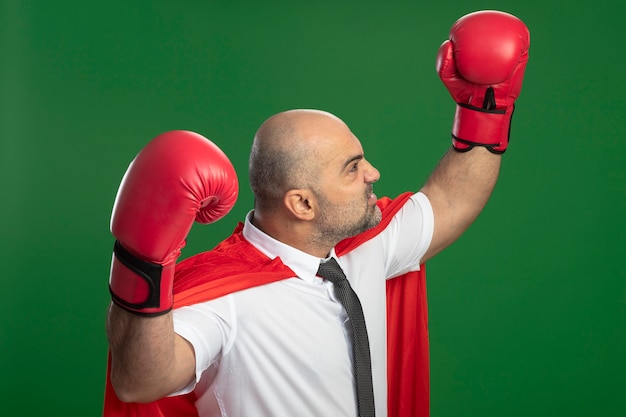 Hombre de negocios de superhéroe enojado en capa roja y guantes de boxeo levantando las manos mostrando fuerza y coraje concepto ganador de pie sobre la pared verde