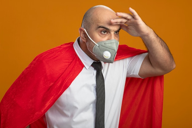 Hombre de negocios de superhéroe en máscara facial protectora y capa roja mirando lejos con las manos sobre la cabeza de pie sobre la pared naranja