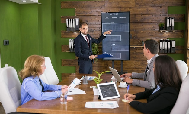 Hombre de negocios en traje apuntando a gráficos en la pantalla del televisor en una reunión con sus colegas. Reunión de liderazgo en sala de conferencias.