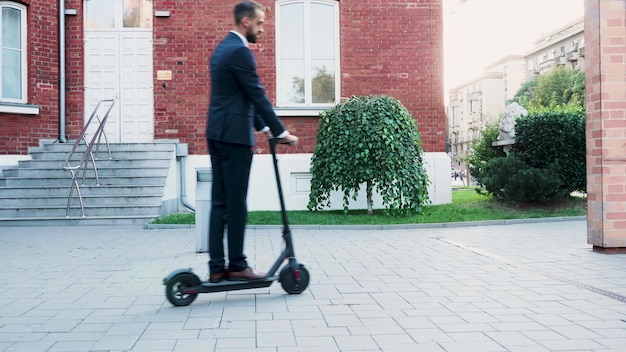 Hombre de negocios en traje diplomático montado en scooter eléctrico frente a la empresa de oficinas de inicio. hombre emprendedor que va a la reunión de negocios. concepto de transporte ecológico