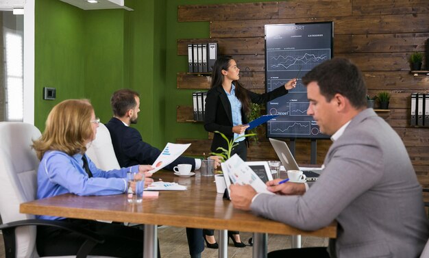 Hombre de negocios en traje escuchando a su colega presentando gráficos en la pantalla del televisor en la sala de conferencias.