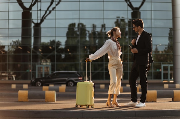 Hombre de negocios en traje negro y atractiva mujer rubia en blusa blanca pantalones beige hablan de buen humor Chica sostiene maleta amarilla y mira a su novio