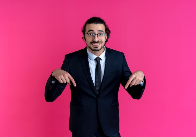 Hombre de negocios en traje negro y gafas apuntando con el dedo hacia abajo mirando al frente sonriendo de pie sobre la pared rosa