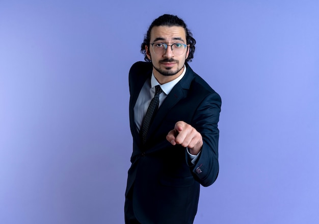 Hombre de negocios en traje negro y gafas apuntando con el dedo hacia el frente mirando confiado de pie sobre la pared azul