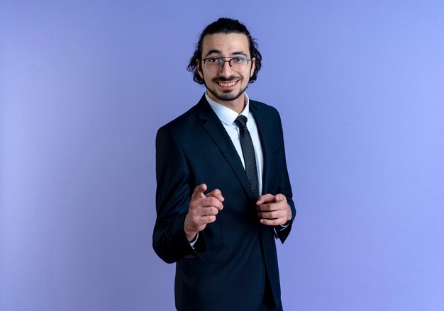 Hombre de negocios en traje negro y gafas apuntando con el dedo hacia el frente sonriendo alegremente de pie sobre la pared azul