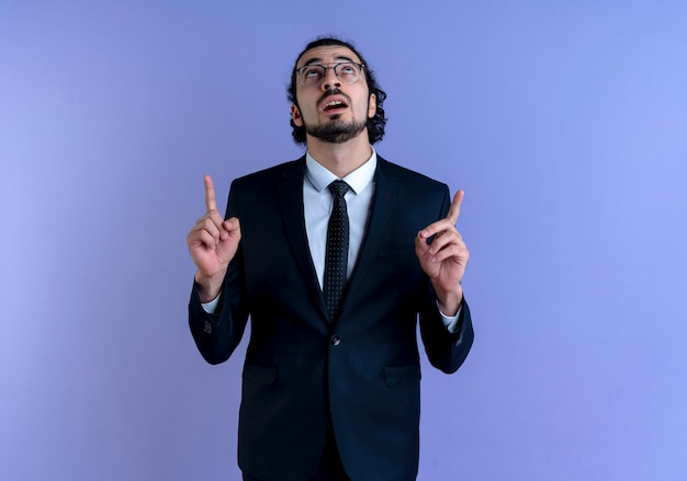 Hombre de negocios en traje negro y gafas apuntando con el dedo índice hacia arriba mirando confiado de pie sobre la pared azul
