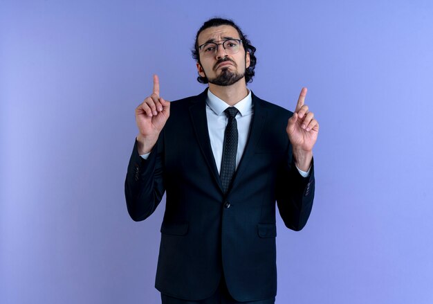 Hombre de negocios en traje negro y gafas apuntando con el dedo índice hacia arriba mirando hacia el frente con expresión triste de pie sobre la pared azul