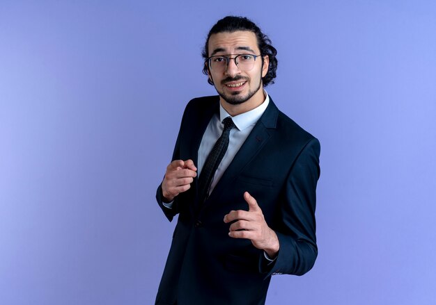 Hombre de negocios en traje negro y gafas apuntando con los dedos índices al frente sonriendo de pie sobre la pared azul