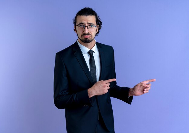 Hombre de negocios en traje negro y gafas apuntando con los dedos índices hacia el lado mirando confundido con expresión triste de pie sobre la pared azul