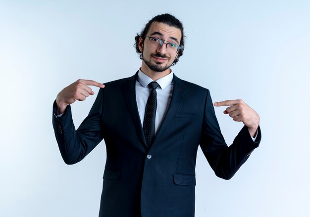 Hombre de negocios en traje negro y gafas apuntando con los dedos índices a sí mismo mirando al frente con una sonrisa en la cara de pie sobre la pared blanca