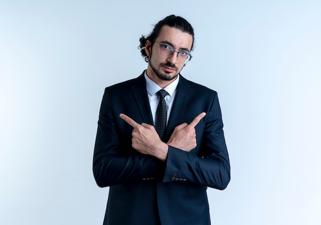 Hombre de negocios en traje negro y gafas apuntando con los dedos hacia los lados mirando confiado de pie sobre la pared blanca