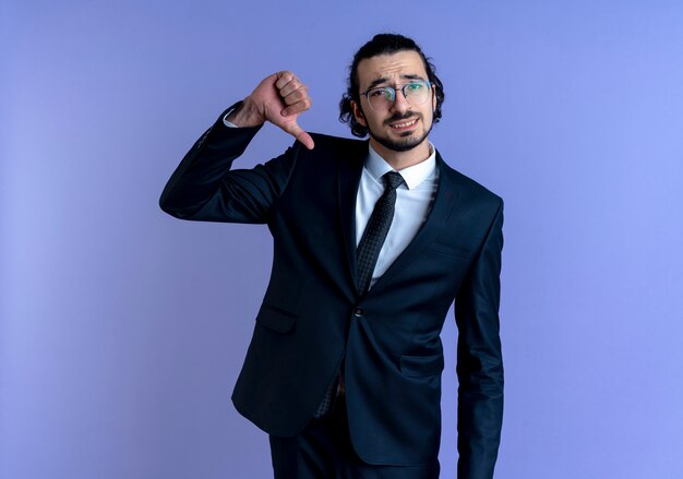 Hombre de negocios en traje negro y gafas apuntando a sí mismo mirando hacia el frente de pie autosuficiente sobre la pared azul