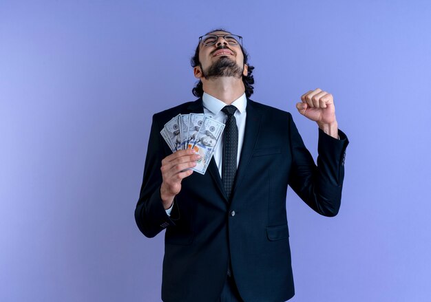 Hombre de negocios en traje negro y gafas con dinero en efectivo feliz y emocionado apretando el puño de pie sobre la pared azul