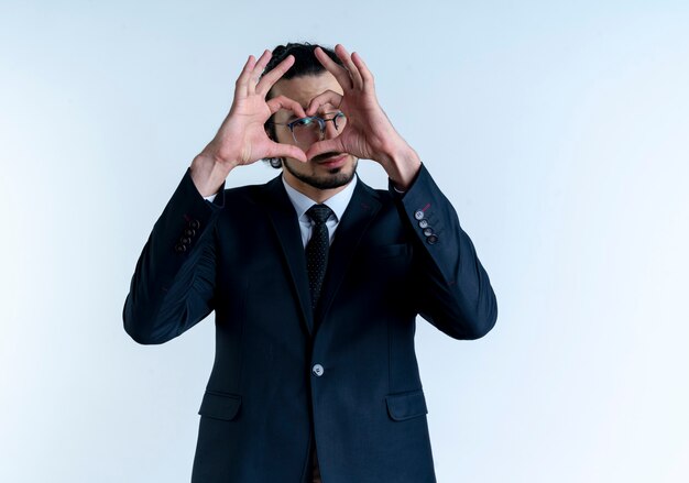 Hombre de negocios en traje negro y gafas haciendo gesto de corazón con los dedos mirando hacia el frente a través de los dedos de pie sobre la pared blanca