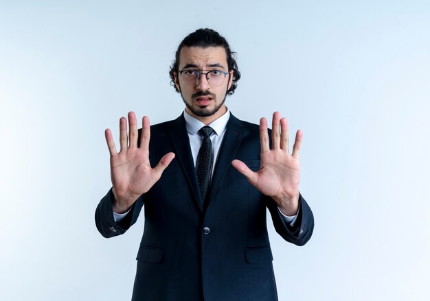 Hombre de negocios en traje negro y gafas haciendo señal de pare con ambas manos mirando hacia el frente con cara seria de pie sobre la pared blanca