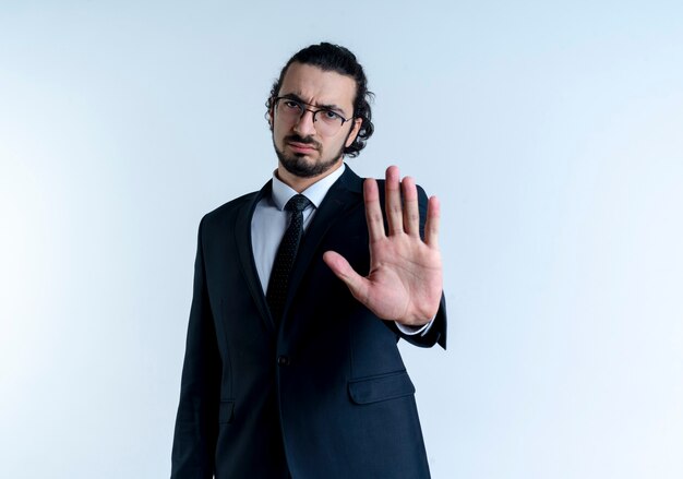 Hombre de negocios en traje negro y gafas haciendo señal de pare con la mano abierta mirando hacia el frente con cara seria de pie sobre la pared blanca