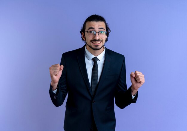 Hombre de negocios en traje negro y gafas mirando al frente apretando los puños feliz y emocionado de pie sobre la pared azul