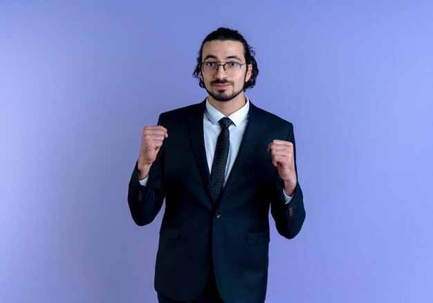 Hombre de negocios en traje negro y gafas mirando al frente apretando los puños feliz y positivo de pie sobre la pared azul