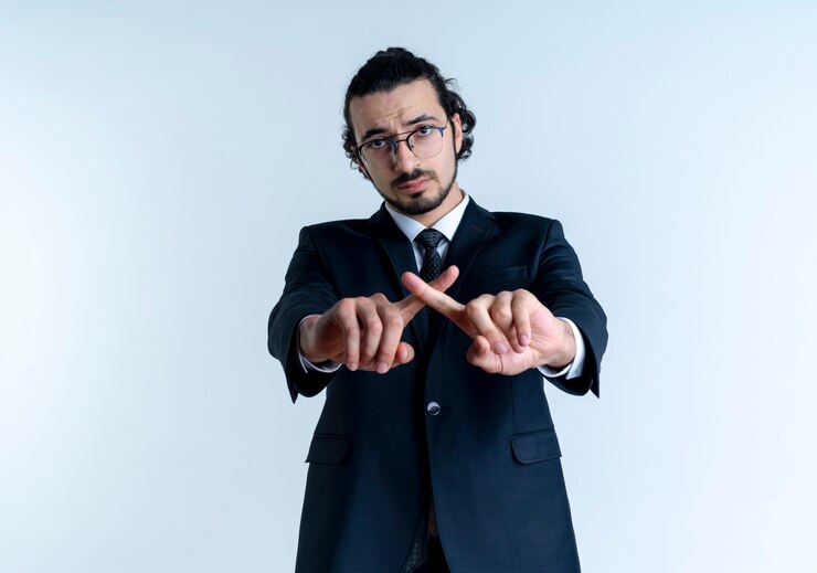 Foto gratuito hombre de negocios en traje negro y gafas mirando al frente con cara seria cruzando los dedos índices haciendo gesto de defensa de pie sobre la pared blanca