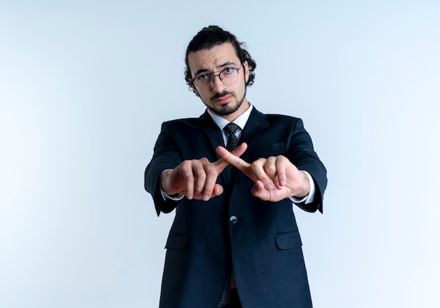 Hombre de negocios en traje negro y gafas mirando al frente con cara seria cruzando los dedos índices haciendo gesto de defensa de pie sobre la pared blanca