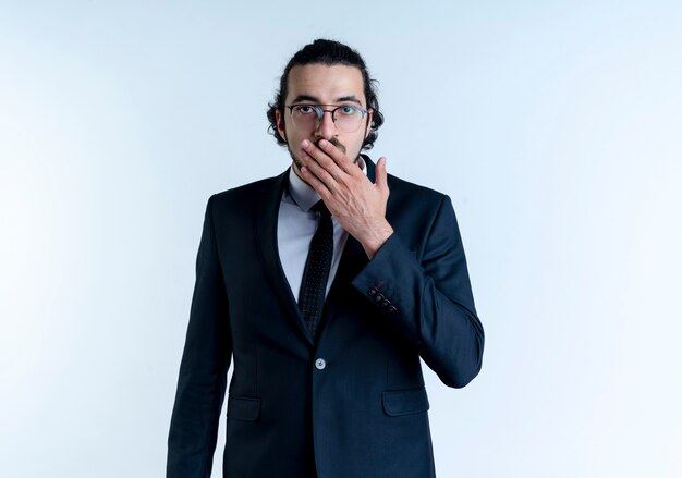 Hombre de negocios en traje negro y gafas mirando al frente conmocionado cubriendo la boca con la mano de pie sobre la pared blanca