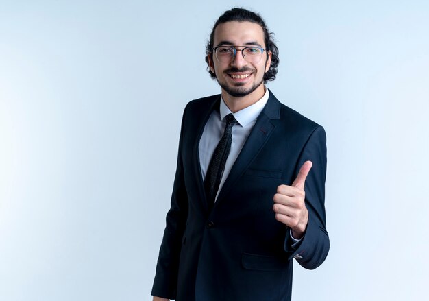 Hombre de negocios en traje negro y gafas mirando al frente con expresión de confianza sonriendo mostrando los pulgares para arriba de pie sobre la pared blanca 2