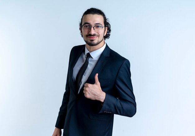 Hombre de negocios en traje negro y gafas mirando al frente con expresión de confianza sonriendo mostrando los pulgares para arriba de pie sobre la pared blanca