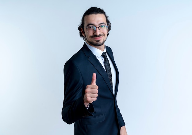 Hombre de negocios en traje negro y gafas mirando al frente con expresión de confianza sonriendo mostrando los pulgares para arriba de pie sobre la pared blanca