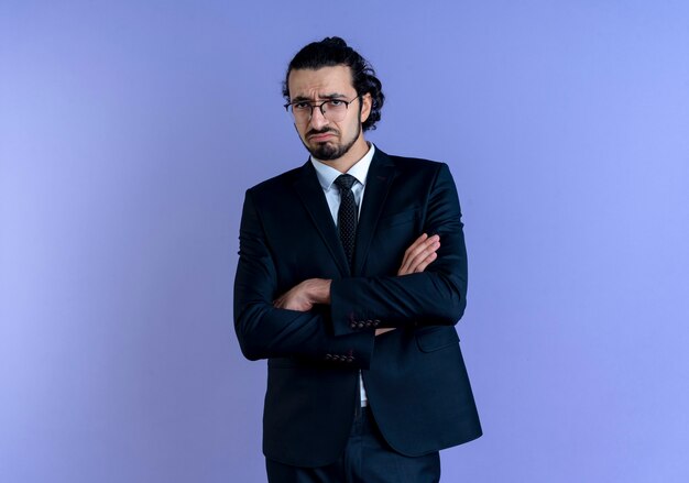 Hombre de negocios en traje negro y gafas mirando al frente con expresión triste en la cara de pie sobre la pared azul